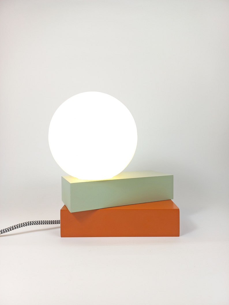 May include: A modern table lamp with a white globe shade and a stacked base of light green and orange rectangular blocks.