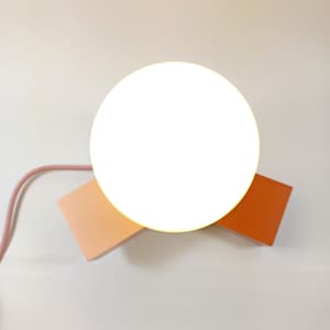 May include: A white round lamp with a pink and orange base. The lamp is on a white surface.
