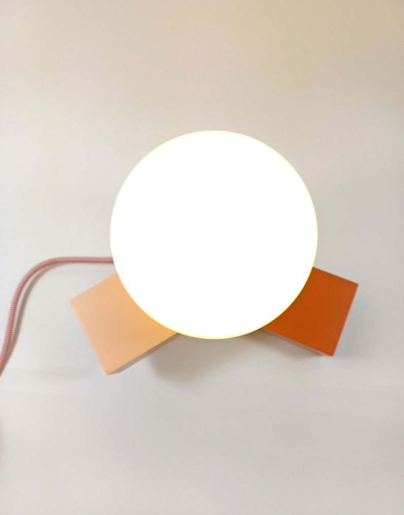 May include: A white round lamp with a pink and orange base. The lamp is on a white surface.