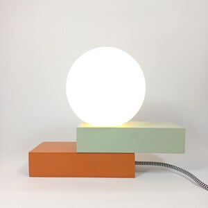May include: A modern table lamp with a white globe shade and a two-tiered base. The top tier is light green and the bottom tier is orange.