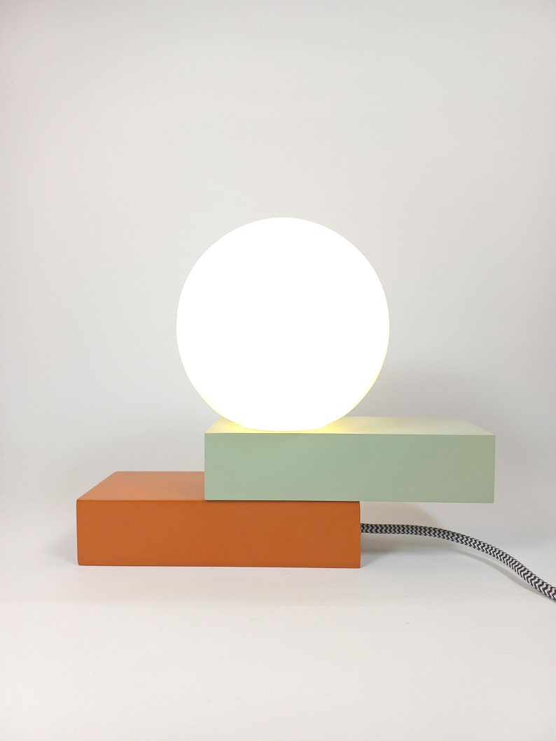May include: A modern table lamp with a white globe shade and a two-tiered base. The top tier is light green and the bottom tier is orange.