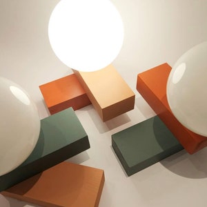 May include: A modern table lamp with two white globe light bulbs. The lamp base is made of four rectangular blocks in shades of orange, green, and brown.