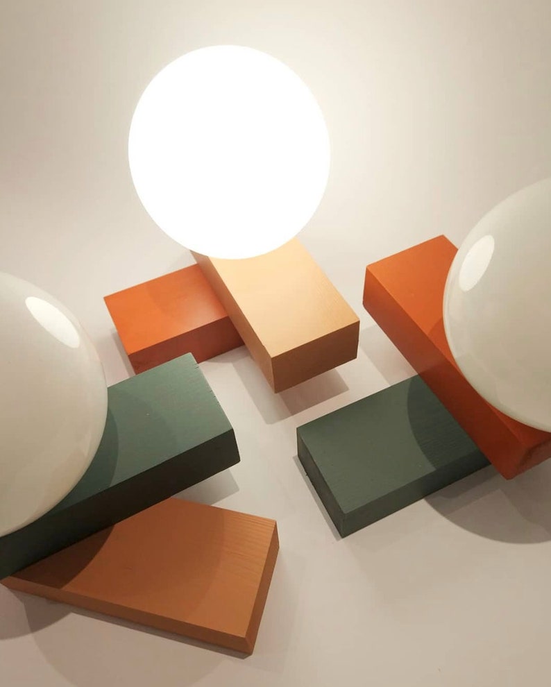May include: A modern table lamp with two white globe light bulbs. The lamp base is made of four rectangular blocks in shades of orange, green, and brown.
