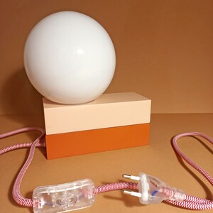 May include: A white globe lamp with a red and white cord and a clear on/off switch. The lamp sits on a two-toned rectangular base with a light pink top and a brown bottom.
