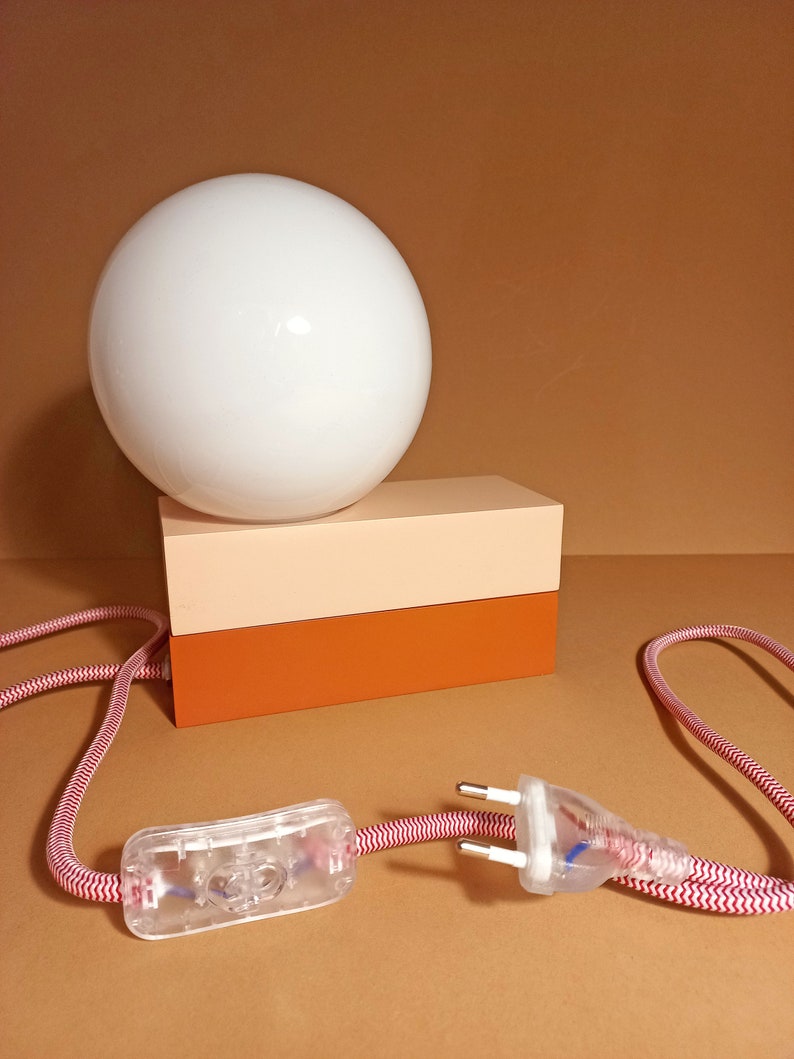 May include: A white globe lamp with a red and white cord and a clear on/off switch. The lamp sits on a two-toned rectangular base with a light pink top and a brown bottom.