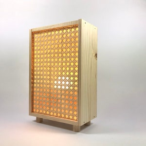 May include: A wooden lamp with a woven cane shade. The lamp is lit and casts a warm glow. The lamp has a rectangular shape and sits on four small legs.