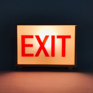 Puede incluir: Un letrero rectangular blanco con marco negro y la palabra "EXIT" en letras rojas.