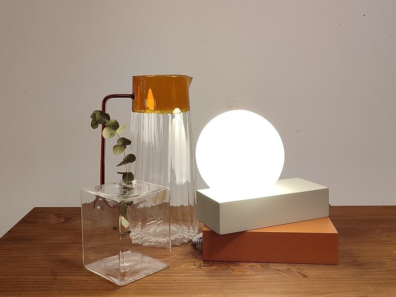 May include: A white globe lamp sits on a light gray rectangular base, which is on top of a brown rectangular base. A clear glass pitcher with a brown top and a clear glass vase with a leafy stem are on a wooden surface.