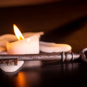 May include: A vintage-style key rests on a melted candle, casting a warm glow. The key is made of metal and has a worn, aged appearance. The candle is white and has a flame burning brightly.