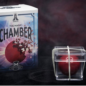 May include: A red ball inside a clear plastic cube with a rubber band around it. The cube is sitting on a dark surface. The box behind the cube has the text "The Magic Chamber" on it.