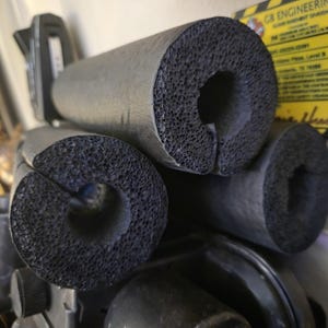 May include: Three cylindrical black foam pipe insulation tubes with a porous, textured surface and a longitudinal slit running the length of each piece. The foam material is dark gray to black with a visible open-cell structure throughout the cross-section.