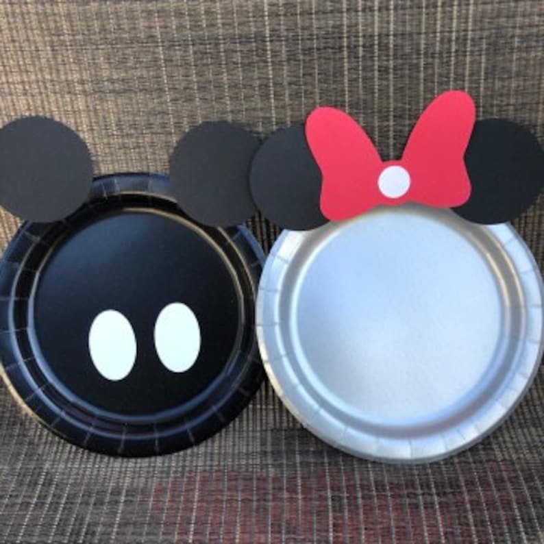 May include: Two round paper plates decorated with character-themed cutouts. One black plate features two black circular ears and two white oval shapes. The other silver plate features two black circular ears and a red bow with a white center dot.