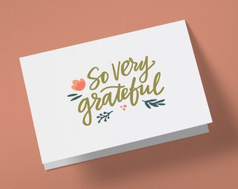 May include: A white card with the text "So very grateful" written in gold script. The text is surrounded by small green leaves and a pink flower.