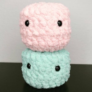 May include: Two crocheted plush toys stacked on top of each other. The top toy is pink and the bottom toy is light blue. Both toys have black button eyes.