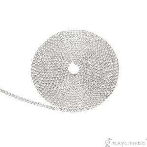May include: A roll of silver rhinestone trim, coiled in a circular shape. The trim is made of small, sparkling rhinestones set closely together, creating a continuous, shimmering band. The trim is unrolled slightly, showing the linear arrangement of the rhinestones.