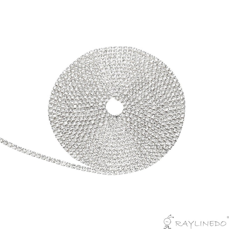 May include: A roll of silver rhinestone trim, coiled in a circular shape. The trim is made of small, sparkling rhinestones set closely together, creating a continuous, shimmering band. The trim is unrolled slightly, showing the linear arrangement of the rhinestones.