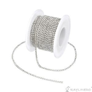 May include: A spool of silver rhinestone chain. The chain is tightly wound around a white plastic spool. The rhinestones are small and closely set, creating a continuous, sparkling line. The chain is unspooled slightly, showing the flexible nature of the chain.