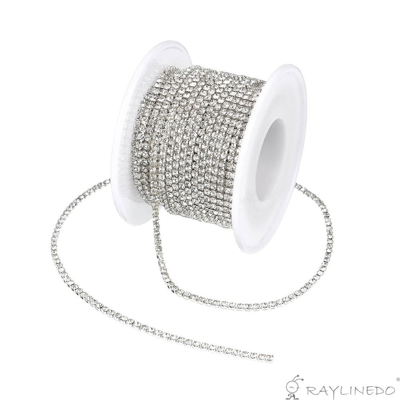May include: A spool of silver rhinestone chain. The chain is tightly wound around a white plastic spool. The rhinestones are small and closely set, creating a continuous, sparkling line. The chain is unspooled slightly, showing the flexible nature of the chain.