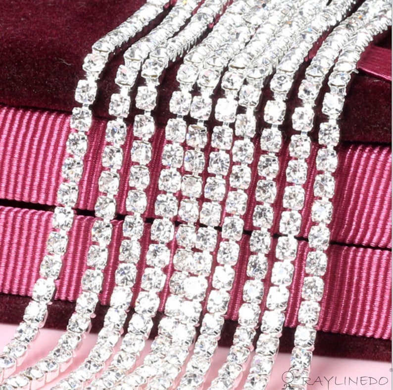 May include: Close-up of multiple strands of sparkling rhinestones. Each strand is composed of square-cut, clear crystals set in a silver-toned metal. The background features a deep burgundy velvet and a striped ribbon.