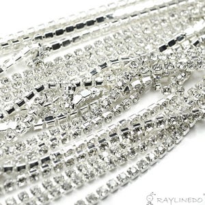 May include: Close-up of multiple strands of silver rhinestone trim. The trim is made of small, square-cut rhinestones set in a silver metal base. The rhinestones are clear and sparkly, reflecting light. The trim is arranged in a layered pattern.