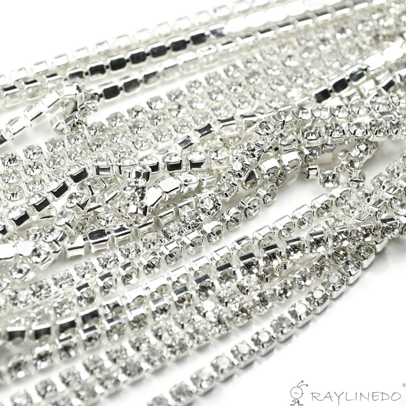 May include: Close-up of multiple strands of silver rhinestone trim. The trim is made of small, square-cut rhinestones set in a silver metal base. The rhinestones are clear and sparkly, reflecting light. The trim is arranged in a layered pattern.