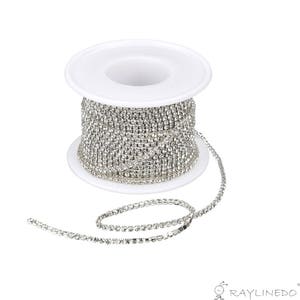 May include: A spool of silver rhinestone chain. The chain is coiled around a white plastic spool, with a length of the chain extending outwards. The rhinestones are small and closely set, creating a sparkling effect. Ideal for jewelry making and crafts.