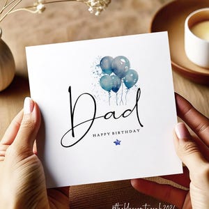 May include: A white birthday card with the word "Dad" in black script, with "Happy Birthday" and blue watercolour balloons. A small blue star is also present. The card is held by hands.