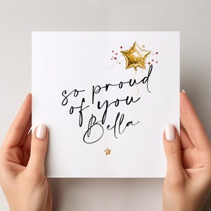 May include: A white card featuring the words "so proud of you Bella" in a stylish script, with a gold star balloon and a small gold star illustration. The card is held by hands with manicured nails.