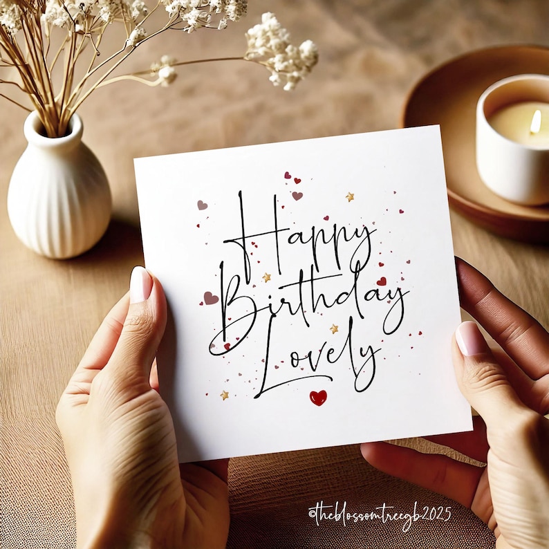 Puede incluir: Una tarjeta de felicitaci&oacute;n blanca con la frase "Happy Birthday Lovely" en escritura elegante, adornada con peque&ntilde;os corazones rojos y estrellas doradas. La tarjeta es sostenida por manos, sugiriendo un regalo reflexivo.