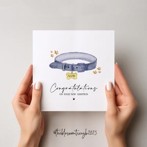 May include: A white card with a watercolor illustration of a blue dog collar with a gold tag that says "Alfie". The card has gold stars and the text "Congratulations on your new addition" with a paw print.