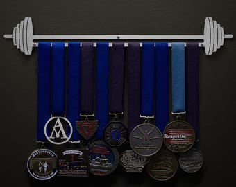 Puede incluir: Un soporte para medallas de metal plateado en forma de barra de pesas con ocho cintas azules y moradas colgando de él. Las cintas sostienen varias medallas con diferentes diseños, incluida una medalla de plata con la letra A, una medalla de plata con un diseño rojo y blanco y una medalla de plata con un diseño azul y blanco.