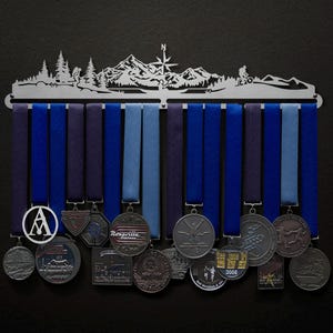 Op de afbeelding: Een zilveren metalen medaillescherm met een bergketenontwerp. De houder heeft 15 haken om medailles aan te hangen. Er hangen verschillende medailles aan de houder, waaronder een zilveren medaille met de tekst "Rosarillo Express".