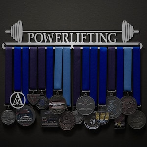 May include: A silver metal medal display rack with the word "POWERLIFTING" printed on it. The rack has 12 hooks for hanging medals. There are several medals hanging on the rack, including a silver medal with the letter "A" on it.