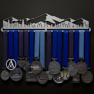 Puede incluir: Un soporte para medallas de metal plateado en forma de cordillera con cintas azules y moradas colgando de él. El soporte tiene una silueta de una cordillera con árboles. Hay varias medallas colgando del soporte, incluida una medalla redonda con la letra "A" en el centro.
