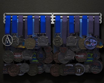 May include: A silver medal rack with blue and purple ribbons holding a collection of silver and black medals. The medals have various designs and logos.
