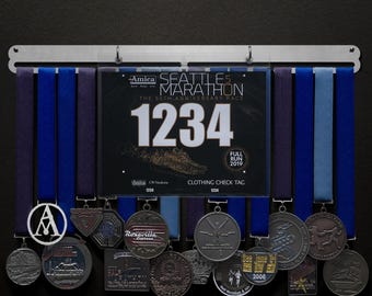 Könnte beinhalten: Ein silberner Medaillenhalter mit blauen und violetten Bändern, die daran hängen. Der Halter hat ein Nummernschild mit der Nummer 1234 und dem Text "Seattle Marathon" darauf. Es hängen mehrere Medaillen am Halter, darunter eine mit dem Buchstaben "A" darauf.
