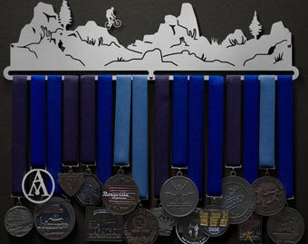 May include: A metal medal display rack with a mountain silhouette design. The rack has 12 hooks for hanging medals and ribbons. The rack is decorated with a mountain scene and a cyclist riding a bike. There are several medals hanging on the rack, including a medal with the text "Rosarilla Express".