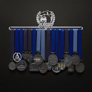 Peut inclure: Un porte-médailles en métal avec un design de tête de bouledogue et le texte "Devil Dog" au-dessus. Le porte-médailles comporte 15 supports de ruban bleus et noirs et contient une variété de médailles argentées et noires avec différents motifs.