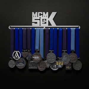 Peut inclure: Un présentoir à médailles en métal argenté avec le texte "MCM 50K" en haut. Le présentoir contient plusieurs médailles avec des rubans bleus. Les médailles ont divers motifs et textes. Le fond est noir.