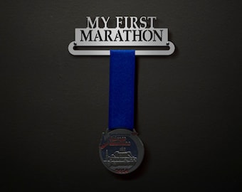 May include: A silver metal medal holder with the text "MY FIRST MARATHON" on it. A blue ribbon is hanging from the holder with a silver medal attached to the end. The medal has a red and white design with the text "Chicago Marathon 2014" on it.