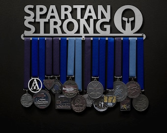 May include: A silver metal medal rack with the words "Spartan Strong" and a Spartan helmet design. The rack has 12 hooks for hanging medals. There are several medals hanging on the rack, including a silver medal with the letter "A" on it.