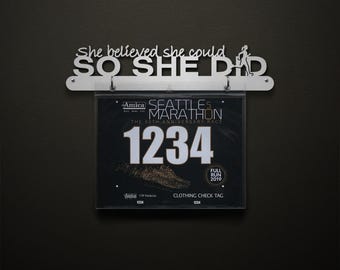 Peut inclure: Un panneau métallique avec l'inscription "She believed she could SO SHE DID" au-dessus d'un support transparent affichant un dossard de course. Le dossard est celui du Marathon de Seattle 5, avec le numéro 1234 et l'année 2019.