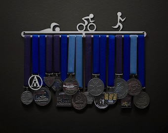 May include: A silver medal rack with a swimmer, cyclist, and runner silhouettes. The rack has 15 hooks and is holding a variety of medals with blue and black ribbons. The medals have different designs, including a logo with the letter A, a logo with the word "Rosarito", and a logo with the year 2006.