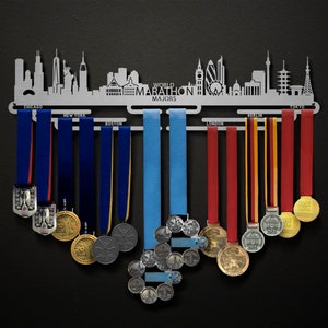 May include: A silver-toned metal medal display rack featuring a skyline silhouette of major cities and the text "WORLD MARATHON MAJORS". The rack includes hooks labelled Chicago, New York, Boston, London, Berlin, and Tokyo, holding various colourful ribbons and medals.