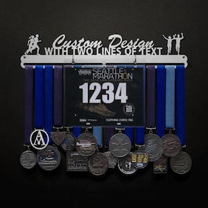 May include: A silver medal display rack with two running figures at either end. The rack has a custom inscription that reads "Custom Design with Two Lines of Text". The rack is holding a variety of medals and ribbons, including a blue ribbon with the text "Seattle Marathon" and a number tag with the number "1234".