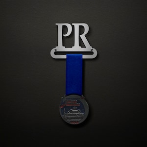 Può includere: Un porta medaglie in argento con le lettere "PR" su di esso. Un nastro blu è appeso al porta medaglie con una medaglia d'argento attaccata all'estremità. La medaglia ha un disegno di un edificio con il testo "Chicago Marathon 2014" su di esso.