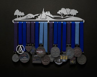 May include: A silver medal display rack featuring a silhouette of trees and a mountain range. The rack has 15 hooks for hanging medals. There are 15 medals hanging on the rack, some with a blue ribbon and some with a red ribbon. The medals are silver and have various designs.