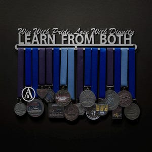 May include: A silver medal rack with the words "Win With Pride, Lose With Dignity LEARN FROM BOTH" hanging on a black wall. The rack has 12 medal hangers and is holding a variety of medals with blue and purple ribbons.
