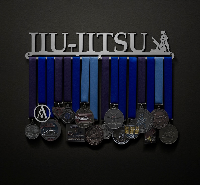 May include: A metal medal display rack with the word "Jiu-Jitsu" in silver lettering. The rack has 12 hooks for hanging medals and ribbons. The rack is decorated with a silver silhouette of a person practicing Jiu-Jitsu. The rack is mounted on a dark gray wall and has a variety of medals hanging from it.