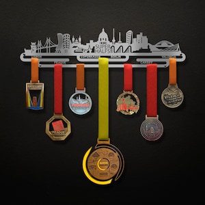 Op de afbeelding: Een metalen medaillescherm in de vorm van een stadsbeeld met de woorden "Super Halfs" in het midden. Het scherm heeft acht haken om medailles op te hangen. Er hangen zeven medailles aan het scherm, waaronder medailles van de halve marathon van Praag, de halve marathon van Valencia en de halve marathon van Cardiff.
